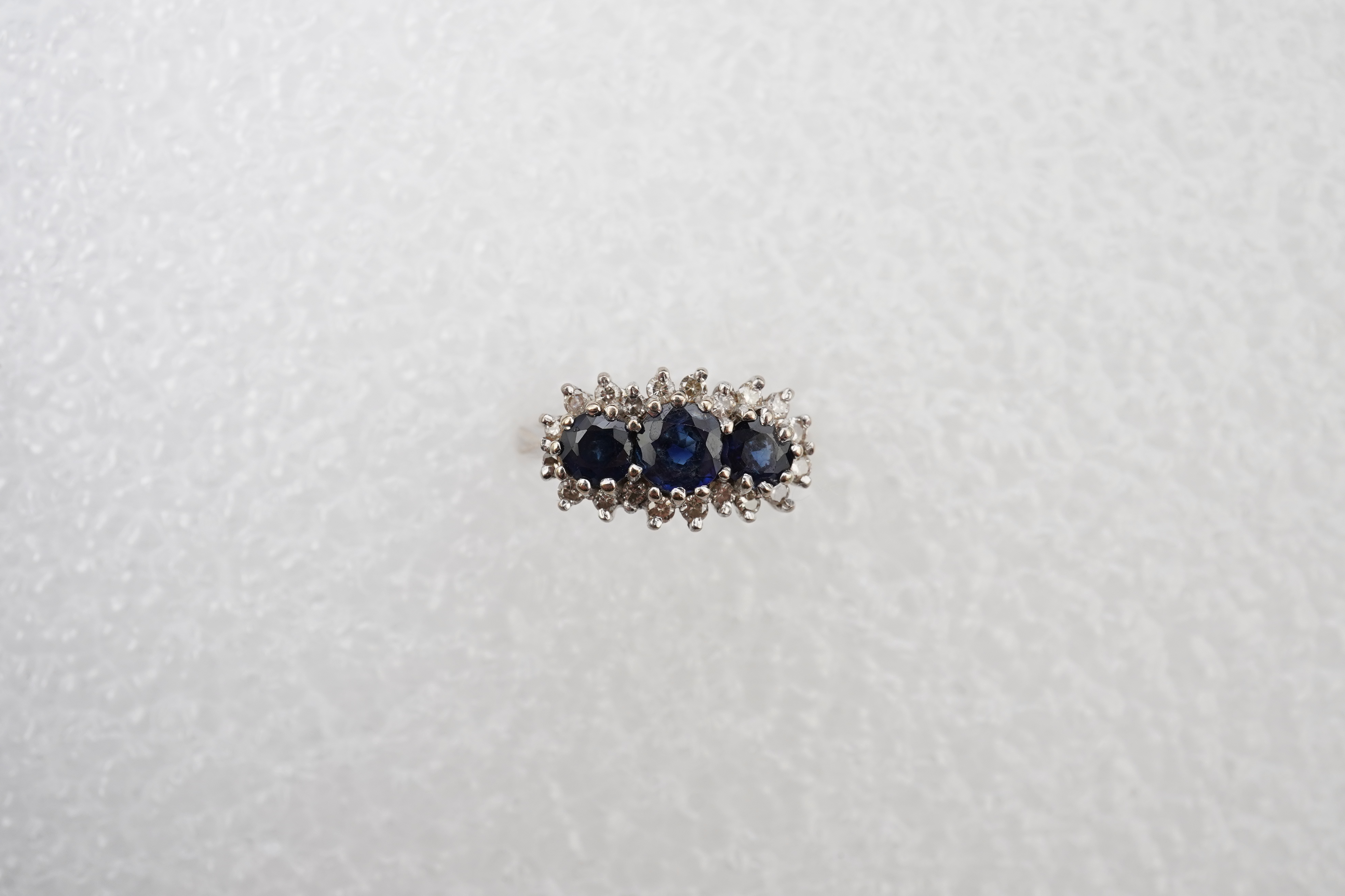 A sapphire and diamond ring, set with three circular-cut and oval sapphires, within a border of brilliant-cut diamonds totalling approximately 0.40 carats, mounted in 18ct white gold, size M1/2, British hallmarks, gross